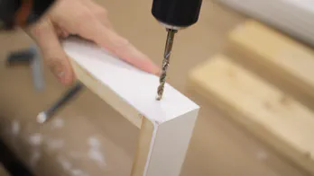 A cordless drill is used to drill a hole in a frame.